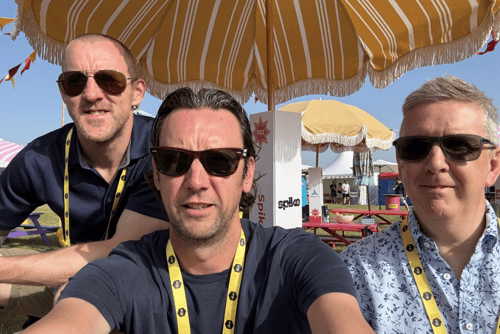 Three white-skinned men wearing sunglasses sitting under an umbrella. They wear lanyards and smart-casual clothing.